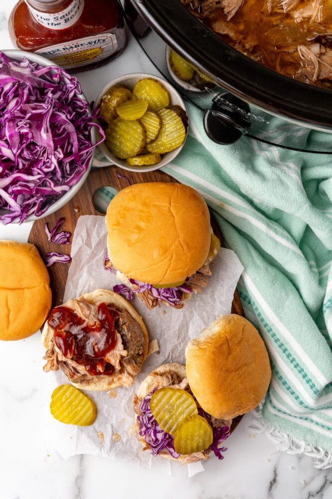 overhead view of crock pot filled with pulled pork, bowl of red cabbage slaw, bowl of butter pickles, bbq sauce, and bbq pulled pork sandwiches