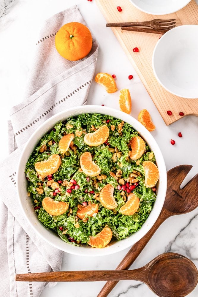 Overhead shot of bowl with kale salad topped with mandarins, pomegranate seeds, and walnuts. It is on top of a white napkin with wood serving spoons and a whole mandarin orange to the side.