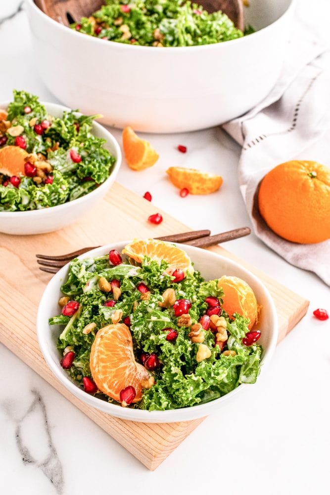 Two small bowls of kale salad on top of a wood serving tray with large bowl of salad in background and and a whole mandarin orange to the side.