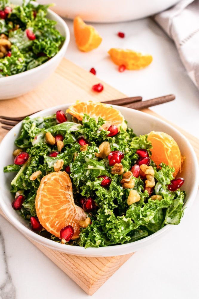 Two small bowls of kale salad on top of a wood serving tray with large bowl of salad in background and and a whole mandarin orange to the side.