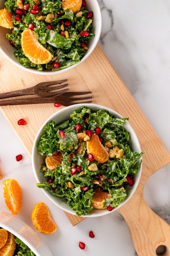 Two small bowls of kale salad on top of a wood serving tray with large bowl of salad in background and and a whole mandarin orange to the side.