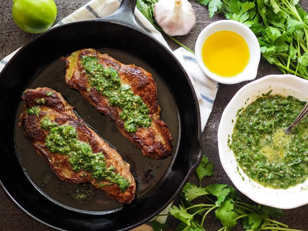 Two NY Strip steaks in a cast iron skillet, seared, and drizzled in a green chimichurri sauce. The to side is a bowl of extra sauce, a lime, and another small bowl of olive oil as well as cilantro and parsley.