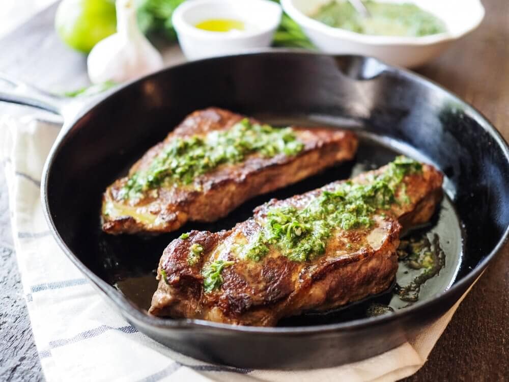 Side view of two NY Strip steaks in a cast iron skillet with the chimichurri sauce and oil on the steaks catching the light fro a nearby window in a delicious way.