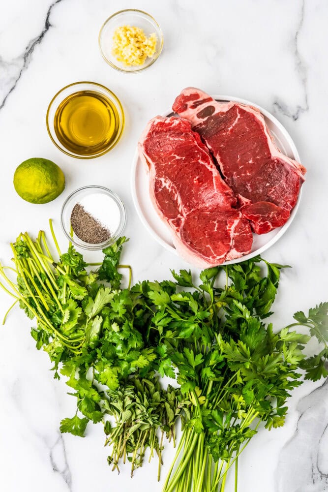 Ingredients to make chimichurri steaks including garlic, olive oil, lime, salt, pepper, cilantro, oregano, parsley, and two NY Strip steaks.