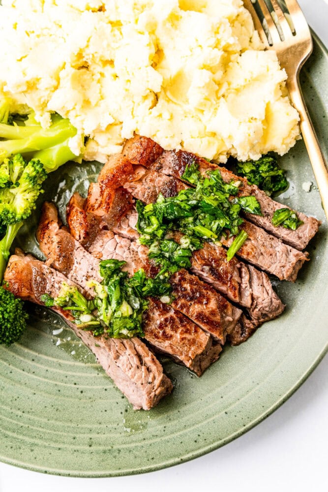 Close up of sliced chimichurri steak on a dinner plate with a side of broccoli and mashed potatoes.