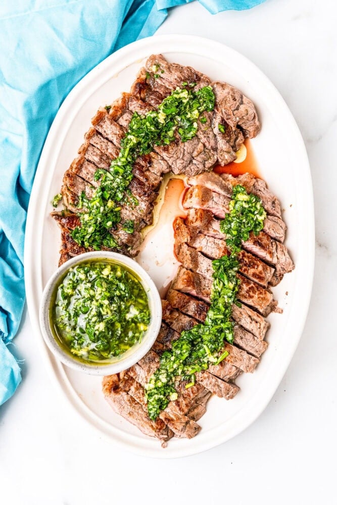 Two NY Strip steaks, sliced and drizzled with chimichurri sauce. Served on a white plate with a turquoise napkin.