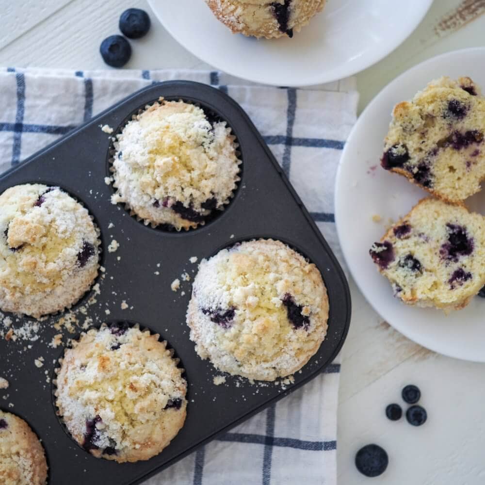lemon blueberry muffins in a muffin tin, placed on a blue and white checkered towel with muffins on white plates to the side.