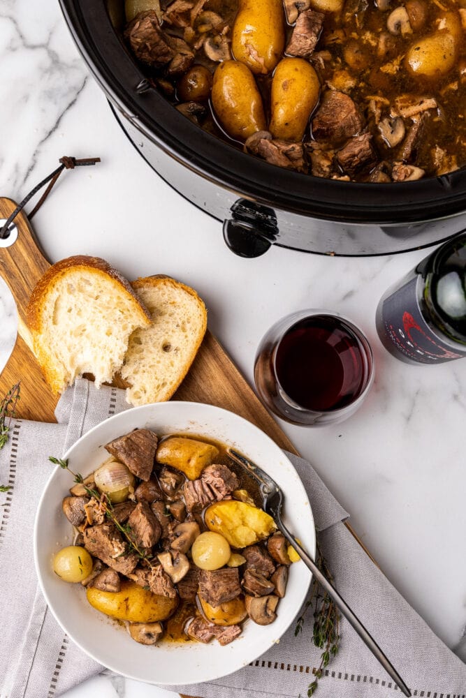 a white bowl of beef bourguignon on a napkin, on a wood board, with French bread, a glass of red wine, and a slow cooker in the background for more servings