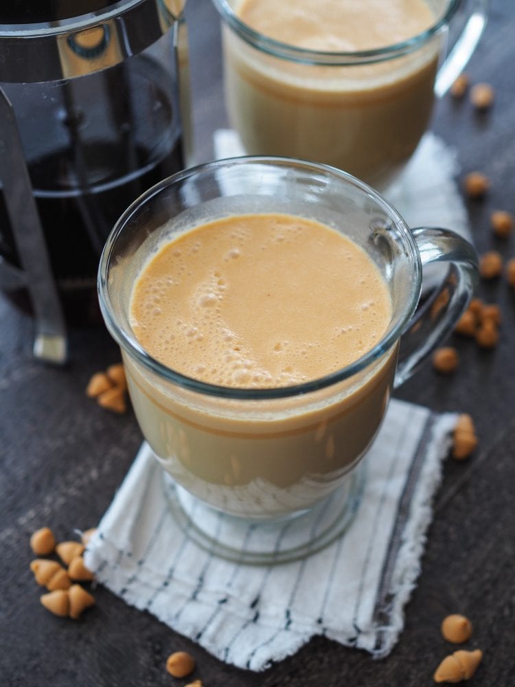 Hot butterbeer in a clear glass mug with a French Press coffee maker and sprinkles of butterscotch chips.