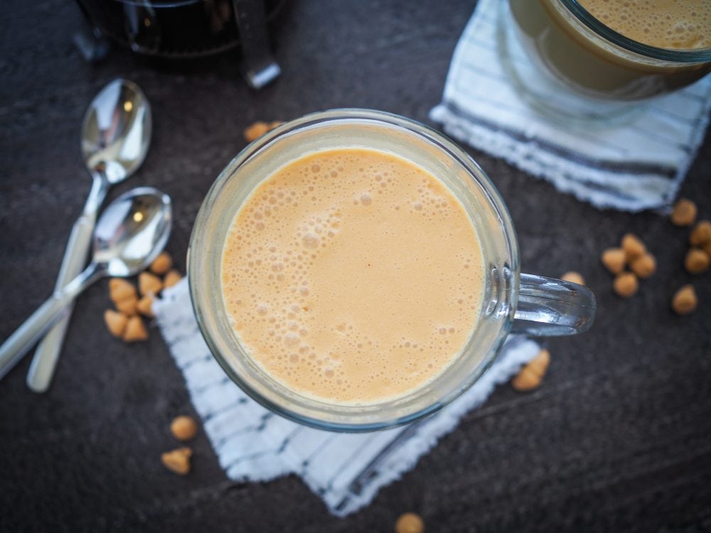 Overhead shot of Hot butterbeer in a clear glass mug with a French Press coffee maker and sprinkles of butterscotch chips.