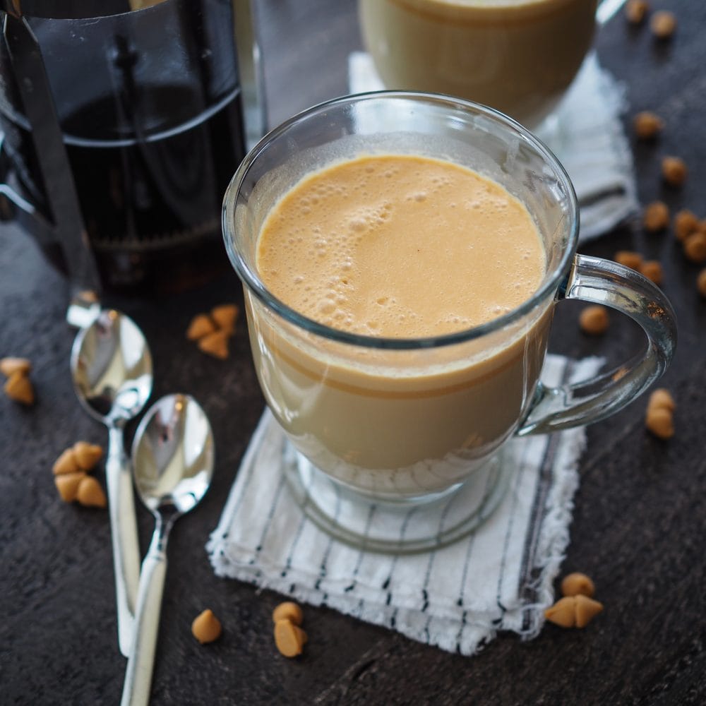 Hot butterbeer in a clear glass mug with a French Press coffee maker and sprinkles of butterscotch chips.