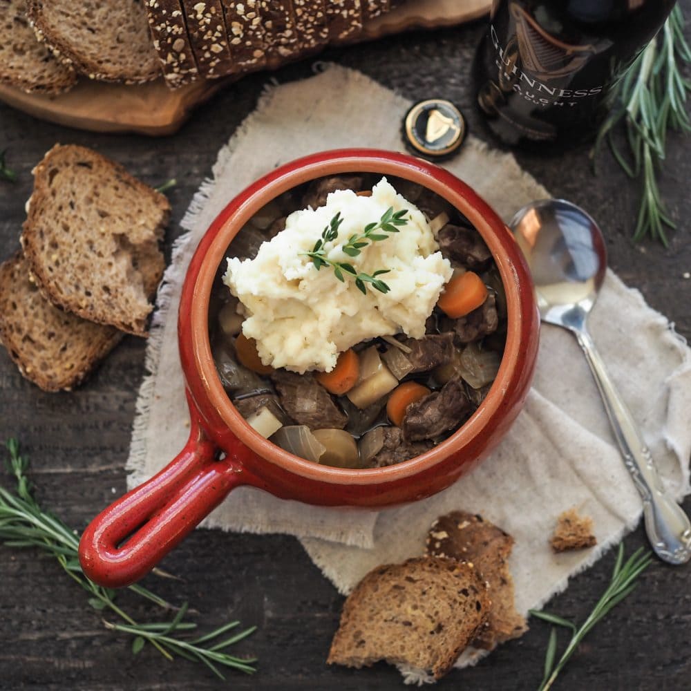 Overhead shot of Guinness Beef Stew in a red crock bowl, topped with mashed potatoes, thyme, and a side of brown soda bread and Guinness.