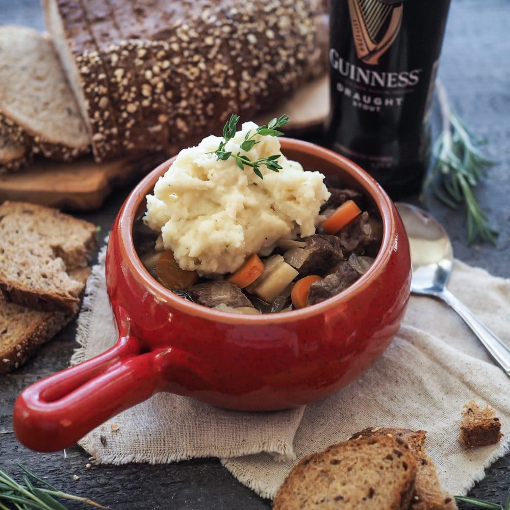 Guinness Beef Stew in a red crock bowl, topped with mashed potatoes, thyme, and a side of brown soda bread and Guinness.