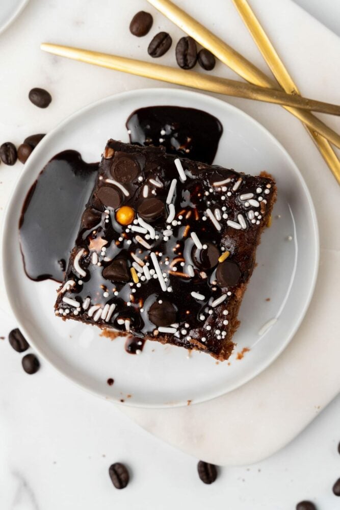 overhead photo of a mocha brownie square on a white plate, covered in white and gold sprinkles. There are coffee beans and gold utensils off to the side.