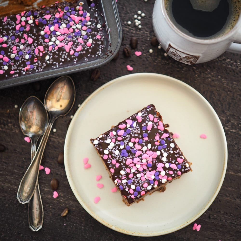 Overhead look at a single square mocha brownie on a plate, with a cup of coffee and pan of brownies to the side. There are pink and purple sprinkles on the brownie.