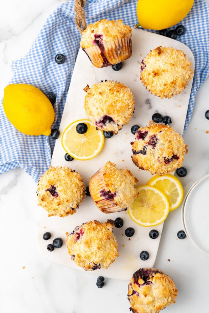 overhead view of fresh baked lemon blueberry muffins on white serving board, surrounded by lemons, lemon slices, blueberries, and blue and white checkered cloth.