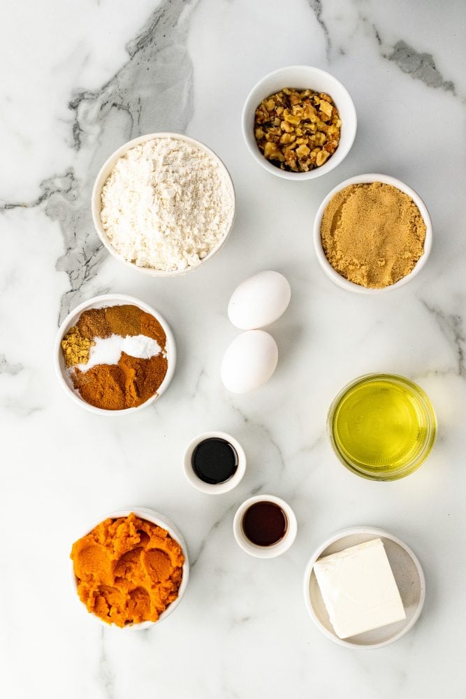 Photo of ingredients for pumpkin cream cheese muffins including flour, brown sugar, spices, eggs, molasses, vanilla, oil, canned pumpkin, cream cheese, and walnuts.