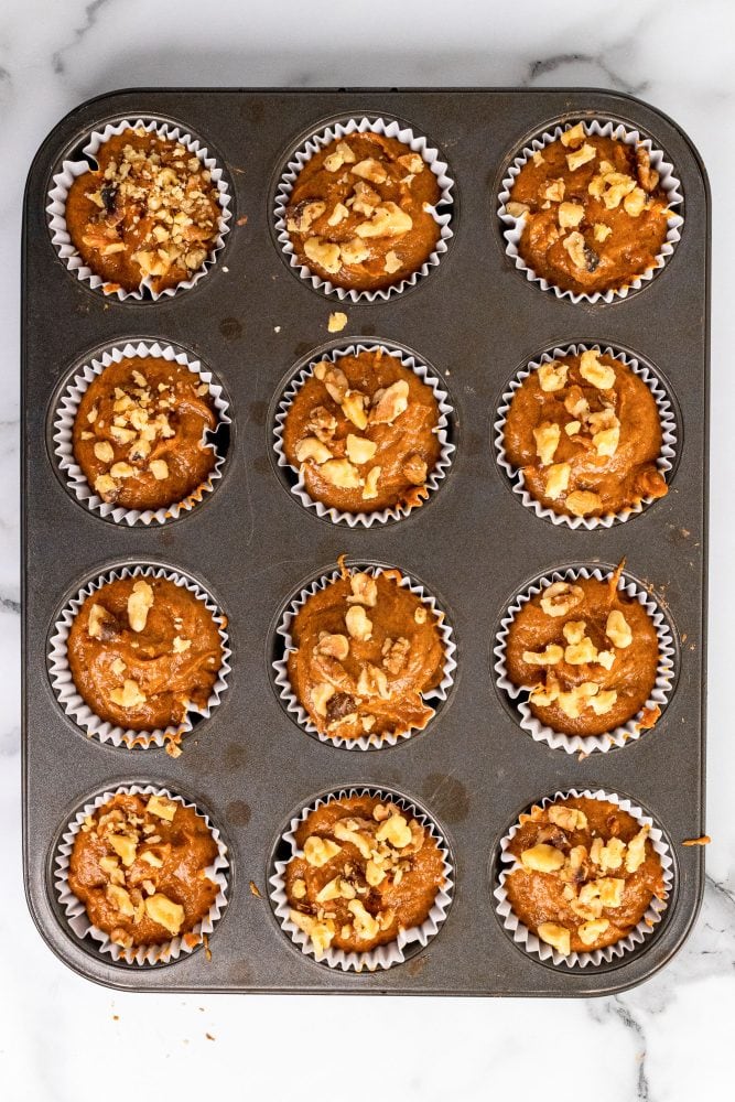 12 un-baked pumpkin cream cheese muffins. The cream cheese as been covered with additional batter and topped with crushed walnuts.