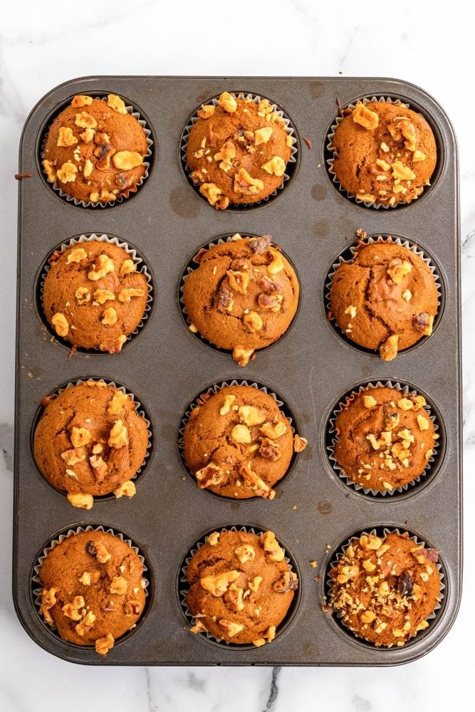 A muffin tin of 12 fresh baked pumpkin cream cheese muffins. The tops are puffed up and rounded and the walnut topping is toasted.