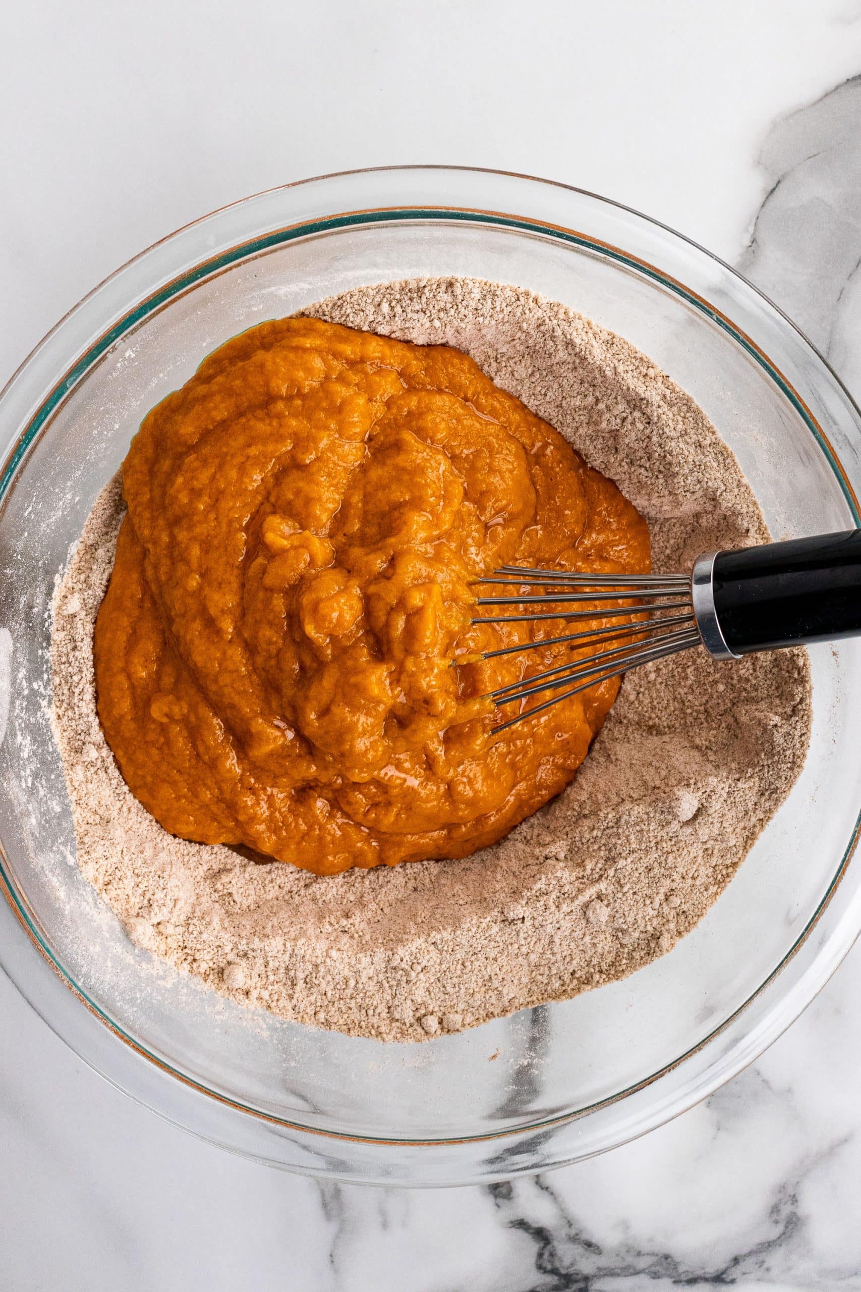 Whisking together wet ingredients and dry ingredients for pumpkin muffins in a clear glass bowl.