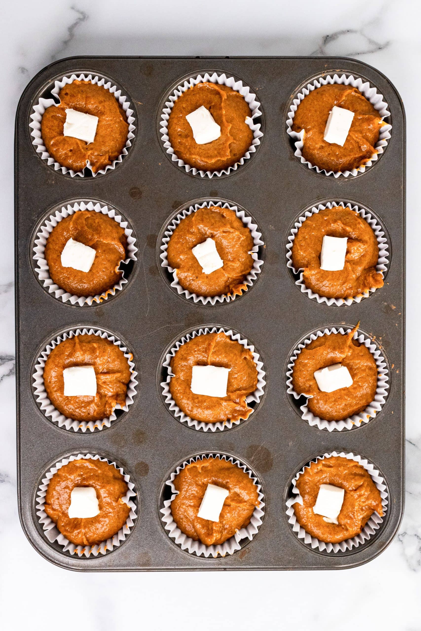 Pumpkin muffin batter in 12 paper cups in a muffin tin with a small cube of cream cheese at the center of each.