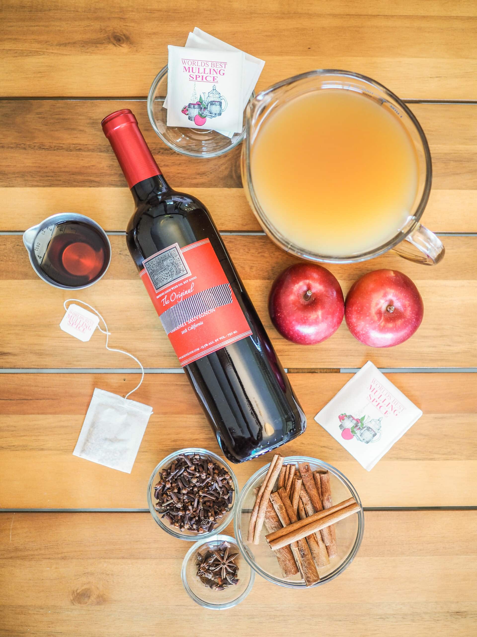 Overhead shot of fall sangria ingredients: shot of whiskey, bottle of red wine, bags of mulling spices, a pitcher of cider, whole apples, cloves, cinnamon sticks, and star anise.