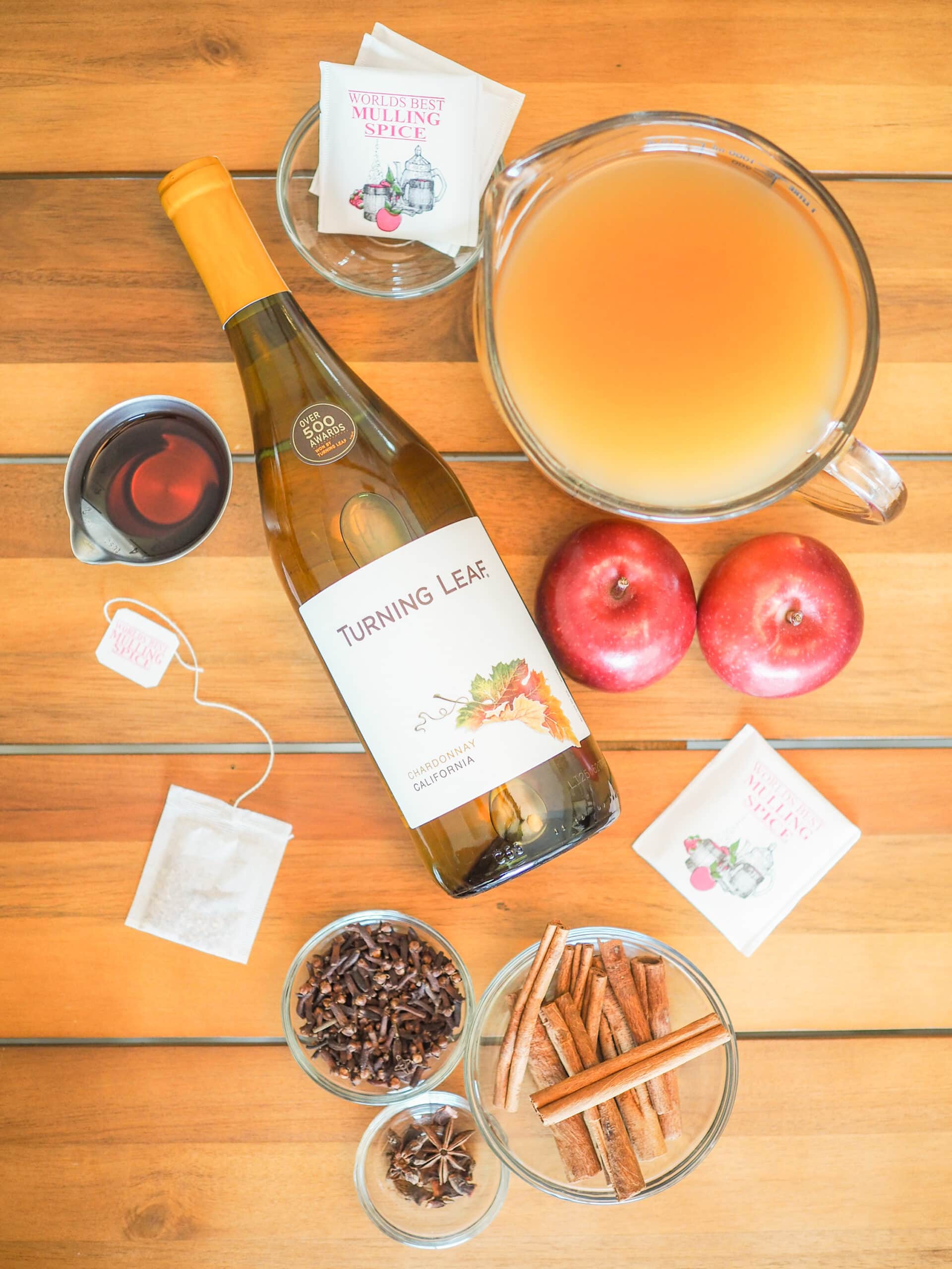 Overhead shot of fall sangria ingredients: shot of whiskey, bottle of white wine, bags of mulling spices, a pitcher of cider, whole apples, cloves, cinnamon sticks, and star anise.