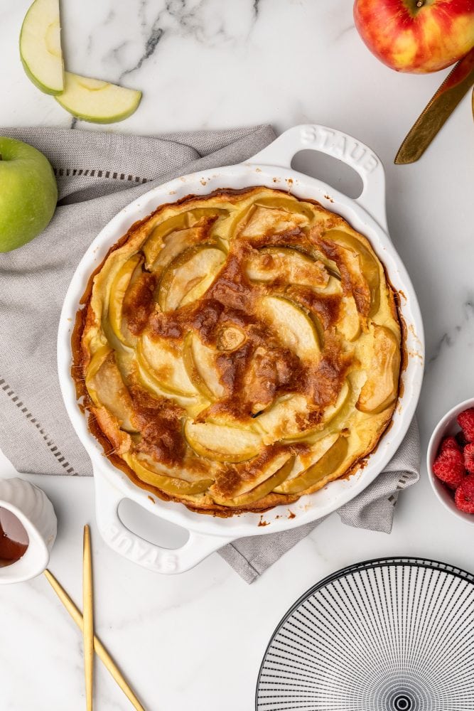 Overhead look at a baked German apple pancake in a white Staub cast iron pie pan on top of a beige napkin with a green apple to the side.