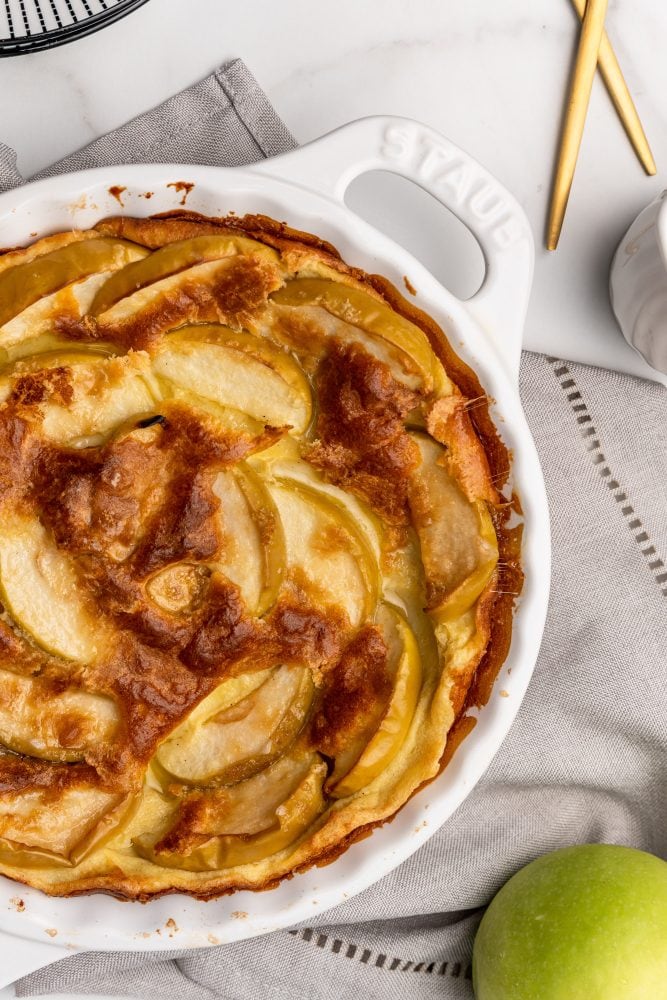 Overhead look at a baked German apple pancake in a white Staub cast iron pie pan on top of a beige napkin with a green apple to the side.