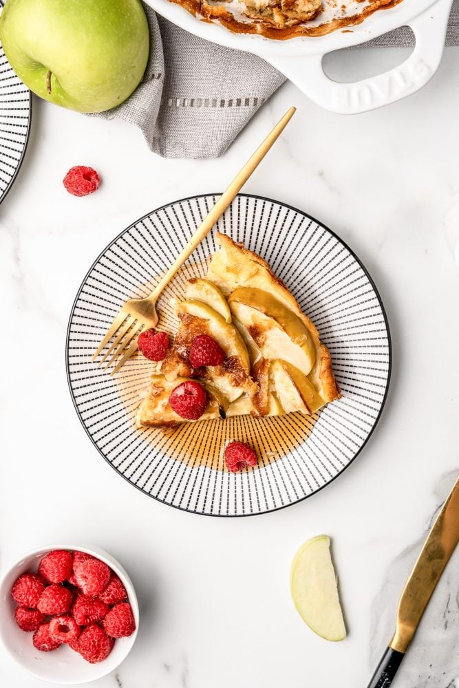 Overhead photo of a black and white striped plate with a slice of apple pancake topped with syrup and fresh raspberries with a gold fork.