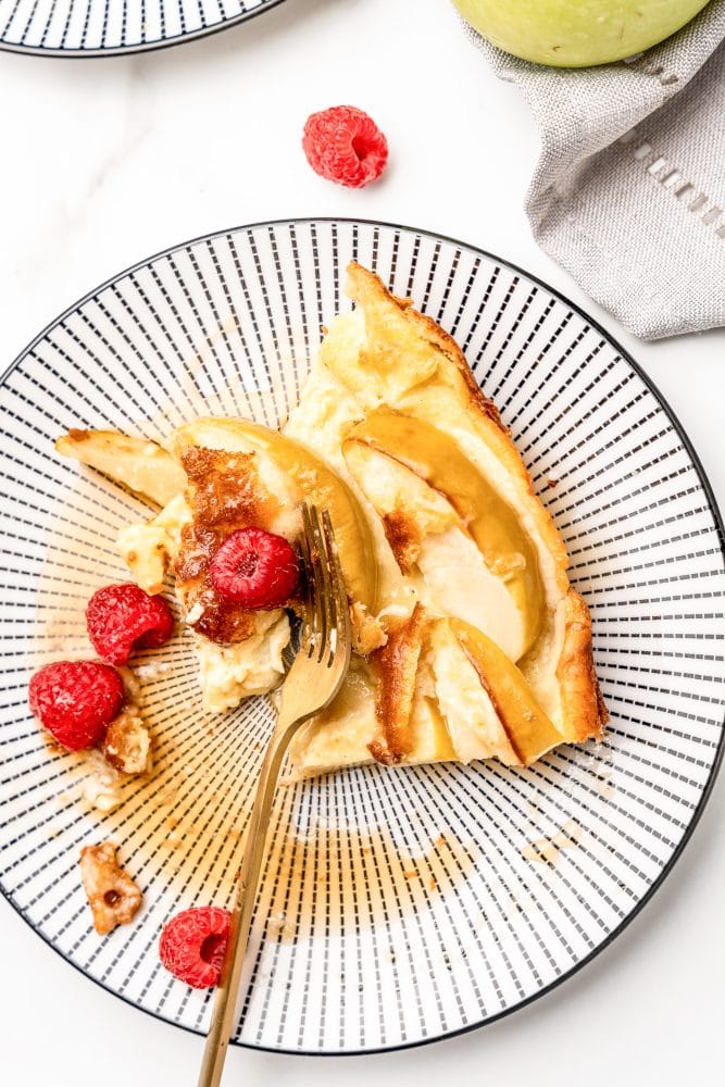 Overhead photo of a black and white striped plate with a slice of German apple pancake topped with syrup and fresh raspberries with a gold fork.