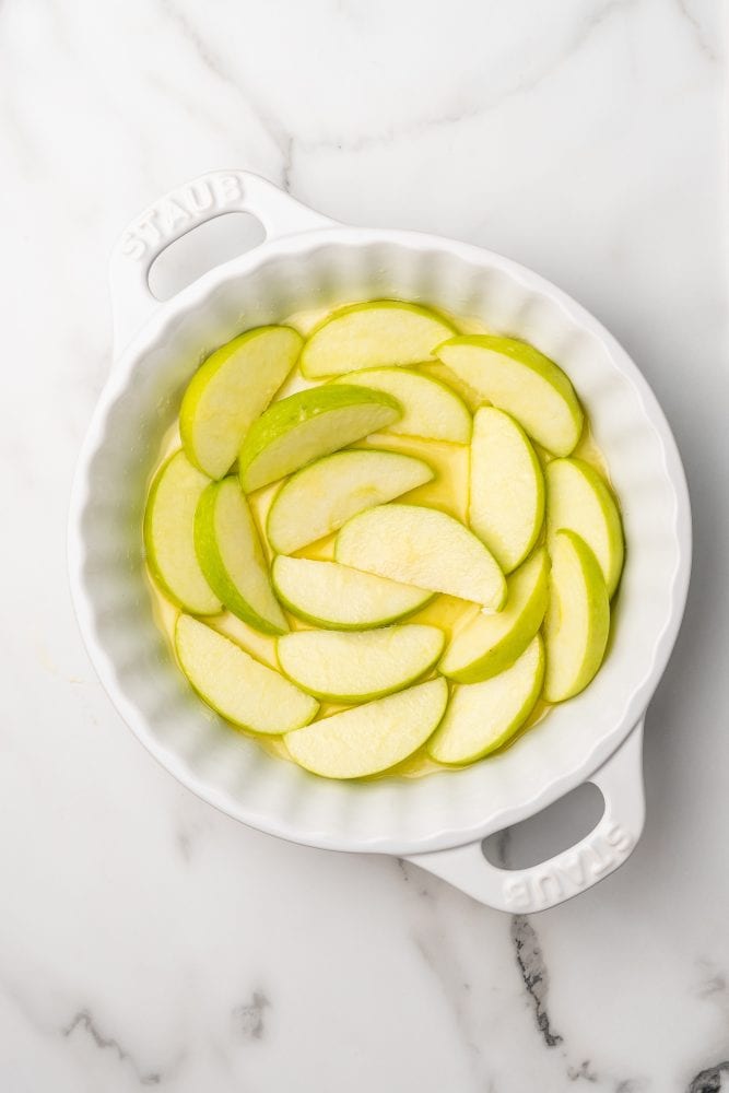 Apple slices on top of melted butter in a white Staub pie dish.