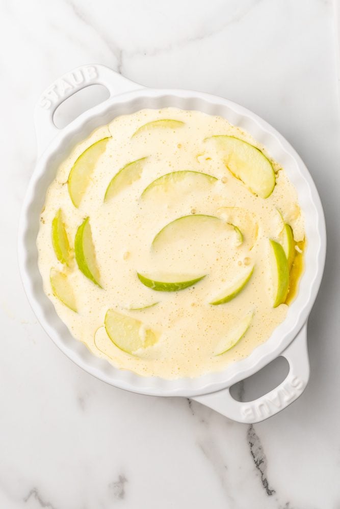 Pancake batter poured on top of apple slices in a white staub pie dish.