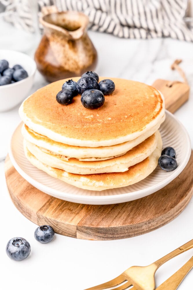 Wood board with white plate topped with stack of four fluffy homemade pancakes and garnished with fresh blueberries.