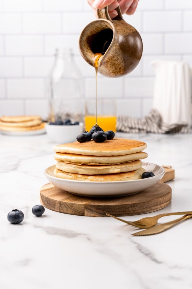 Wood board with white plate topped with stack of four fluffy homemade pancakes and garnished with fresh blueberries. A small earthenware container is slowly pouring syrup on top.