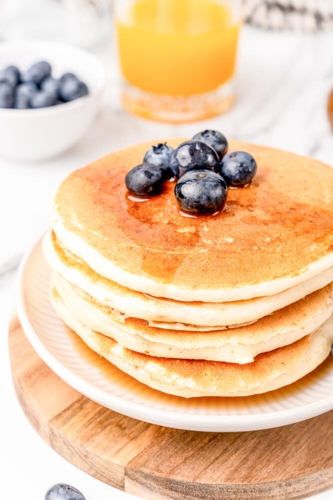 Stack of four pancakes on a white plate sitting on a wood board. The pancakes are topped with blueberries and syrup. And there's a bowl of orange juice and fresh berries in the background.