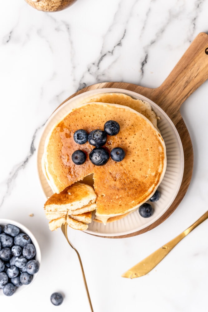 Overhead shot of stack of pancakes with a forkful of three fluffy pancake bites. There's a bowl of blueberries off to the side.