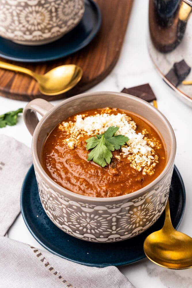 bowl of spicy tomato soup in a grey bowl, resting on a blue plate with gold spoon next to it. It's garnished on top with cheese and parsley.