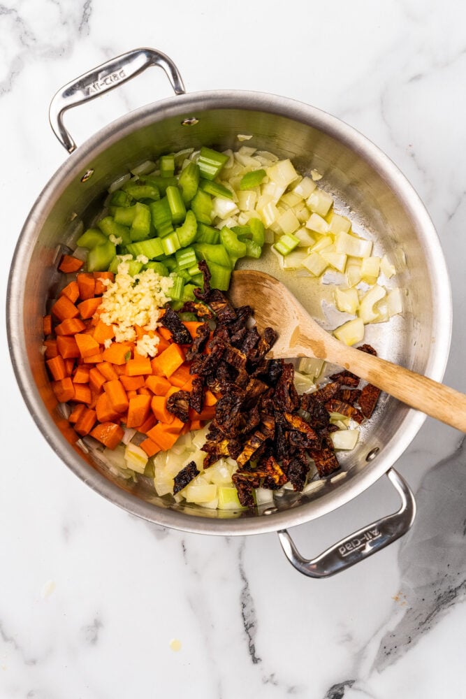 Adding in other vegetables to soup pot including carrots, celery, sun dried tomatoes, and garlic.