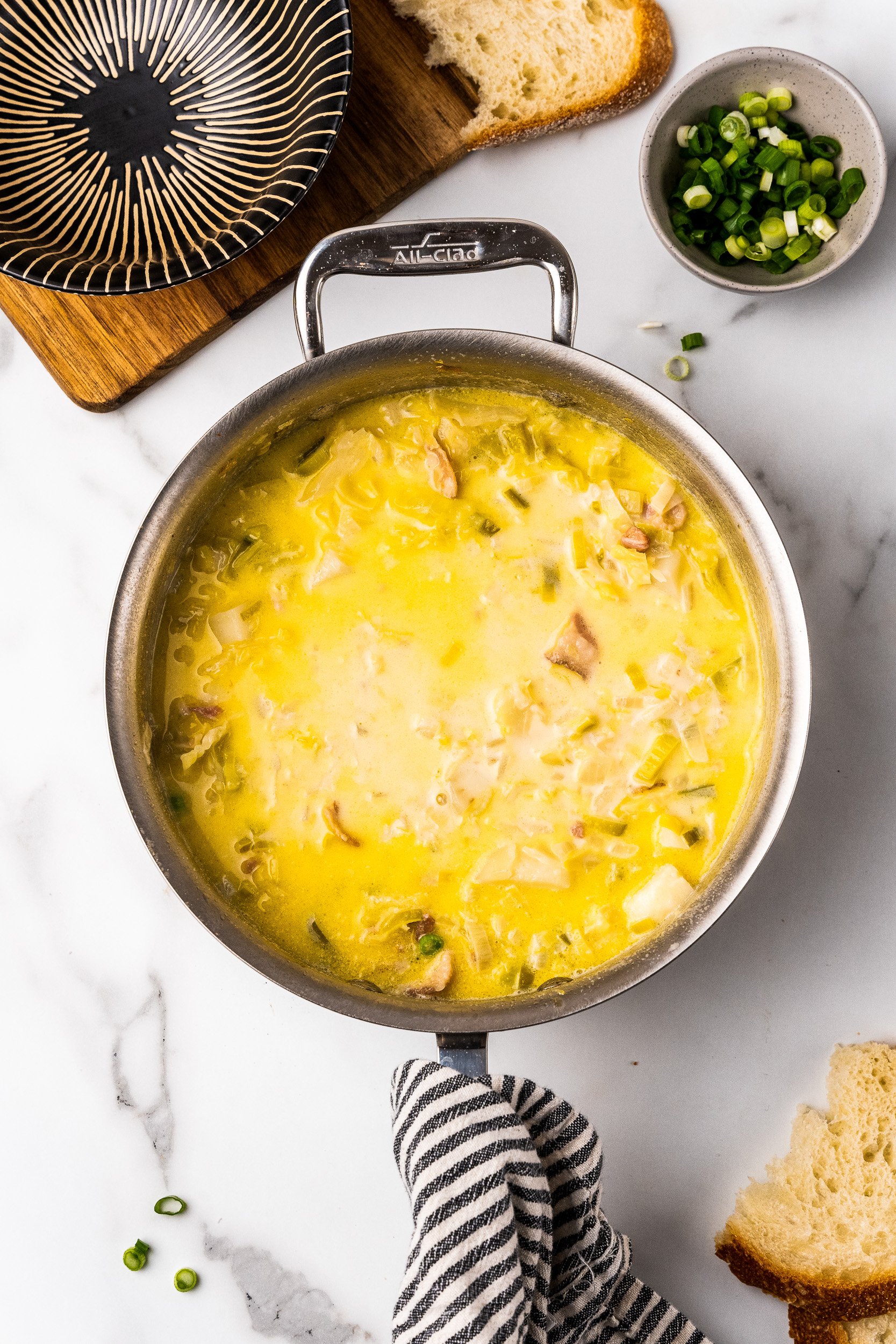 Colcannon soup in a stainless steel All Clad soup pan. There's a black and cream striped bowl to the side waiting for a serving, as well as torn bread and a small bowl of green onions for garnish.