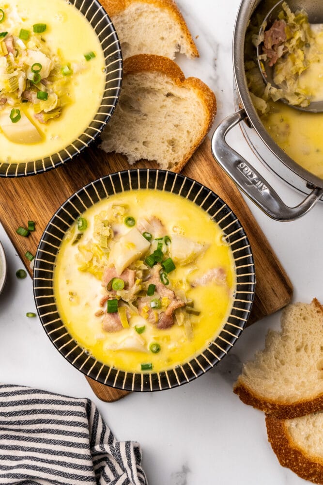 Comforting bowl of Irish colcannon soup. There are bits of bacon, onions, cabbage, and potatoes resting in a creamy buttery broth in a black striped bowl. There is bread on a wood board to the side, and the stainless steel soup pan ready for more to be served sitting next to the bowl.