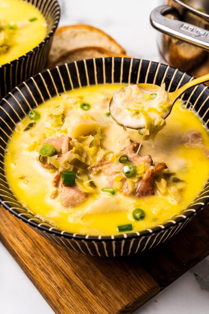 Comforting bowl of Irish colcannon soup. There are bits of bacon, onions, cabbage, and potatoes resting in a creamy buttery broth in a black striped bowl with a golden spoon lifting out a bite. 