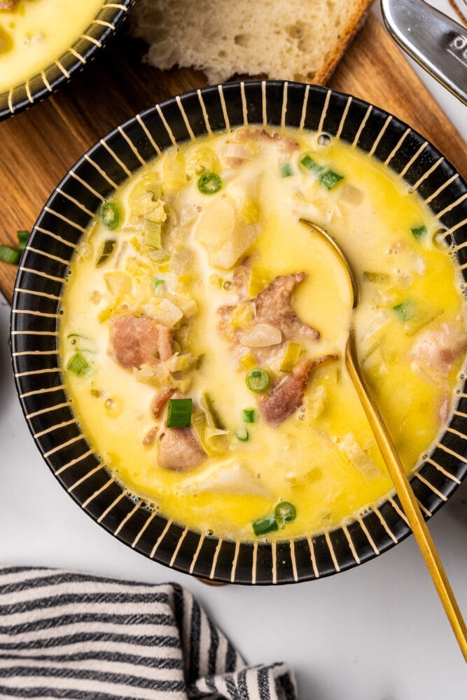 Comforting bowl of Irish colcannon soup. There are bits of bacon, onions, cabbage, and potatoes resting in a creamy buttery broth in a black striped bowl with a golden spoon.