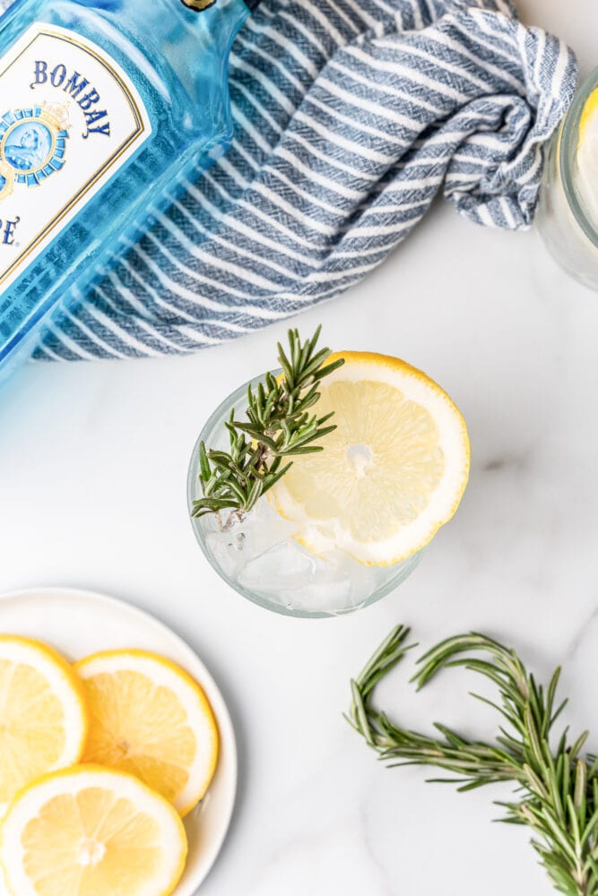 overhead of lemon rosemary gin fizz with sprig of fresh rosemary, slice of lemon, plate of sliced lemons to the side, and blue towel and bottle of Bombay gin in background.