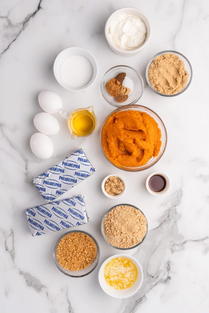 Ingredients for easy pumpkin cheesecake recipe with bourbon cream including: sour cream, white sugar, pumpkin pie spices, brown sugar, whiskey, pumpkin, cream cheese, vanilla, eggs, crushed graham crackers, crushed ginger snaps, and melted butter.