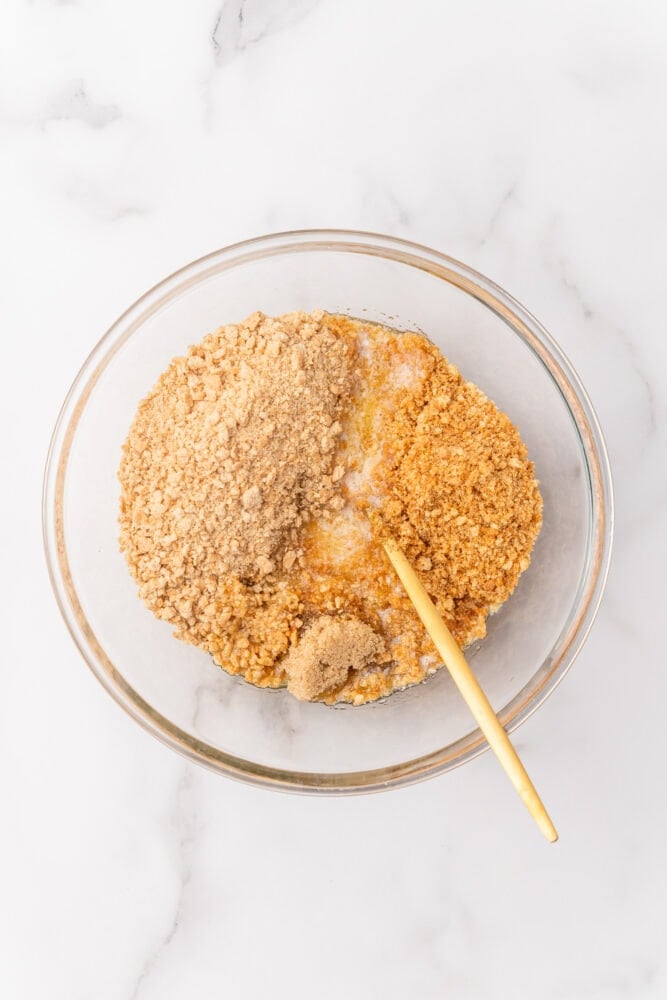 Crushed ginger snaps, crushed graham crackers, and melted butter in a bowl with a gold spoon ready to be mixed together.