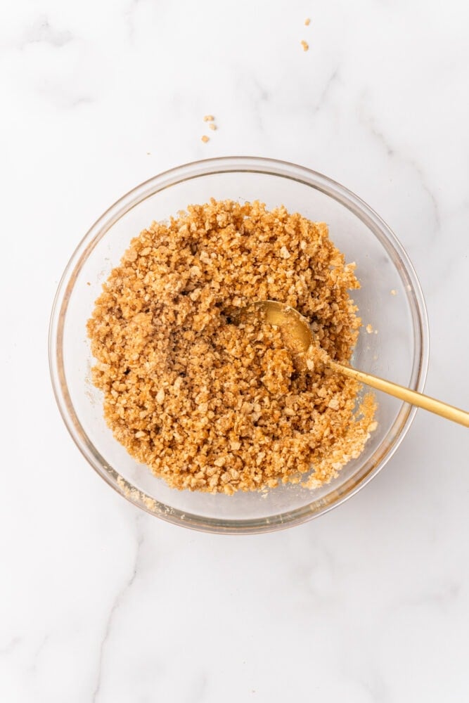 Crushed ginger snaps, crushed graham crackers, and melted butter in a bowl with a gold spoon all blended together.