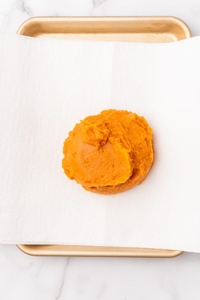 Canned pumpkin on top of a layer of white paper towels on a gold baking sheet.