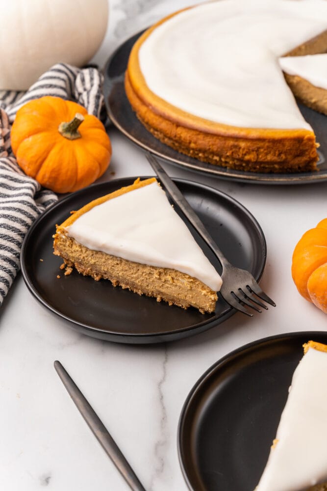 Slice of pumpkin cheesecake with bourbon cream on a black plate, the whole cheesecake and mini decorative pumpkins in the background