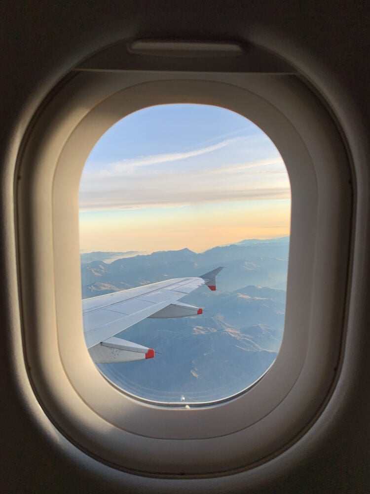 Photo looking out of oval airplane window at tip of the wing of a plane over mountains.